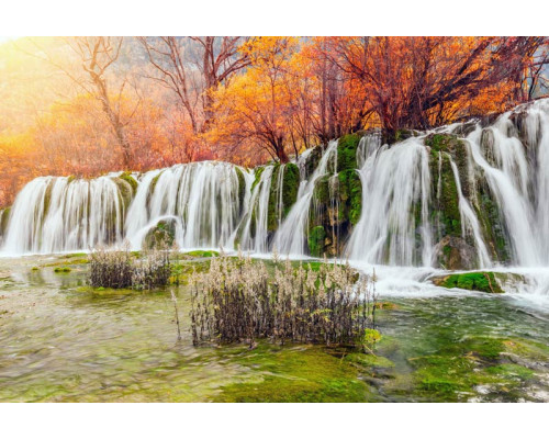 Fotobehang van een waterval die door een herfstlandschap stroomt, met kleurrijke bomen en bladeren eromheen.