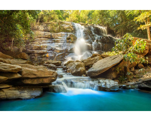 Fotobehang van een zonnige waterval die door rotsen stroomt, met een helderblauwe lucht en een stralende zon.