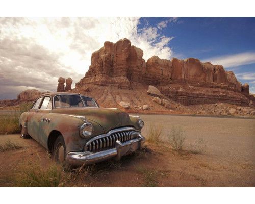 Fotobehang van een oude auto die in een woestijnlandschap tussen rotsen staat, met een helderblauwe lucht.