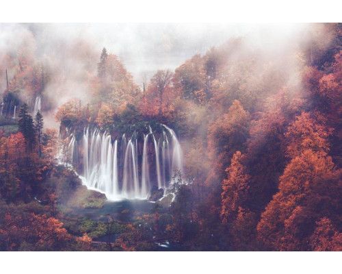 Fotobehang van een waterval die door een mistig herfstbos stroomt, omringd door bergen en bomen.