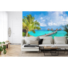 Een fotobehang met een tropische vakantiesfeer met palmbomen op een strand en een blauwe zee, afgebeeld in een huiskamer.