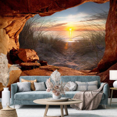 Een fotobehang van een zonsondergang vanuit een grot met uitzicht op de duinen en zee in vliesbehangstijl voor een woonkamer.