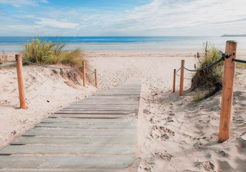 Zandpad dat leidt naar een afgelegen strand met helderblauwe zee en duinen op de achtergrond, fotobehang dat rust en vrijheid uitstraalt.